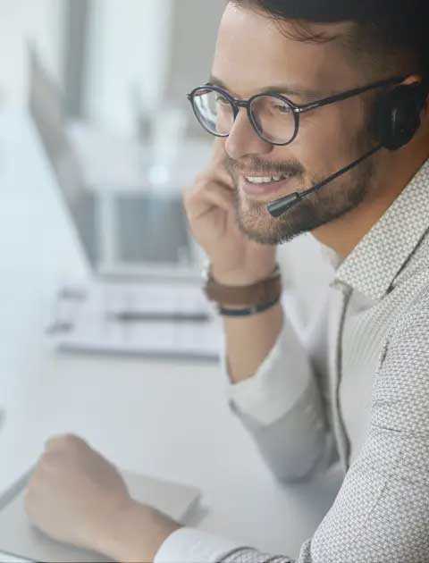 Man with glasses sitting at a desk talking into a headset.